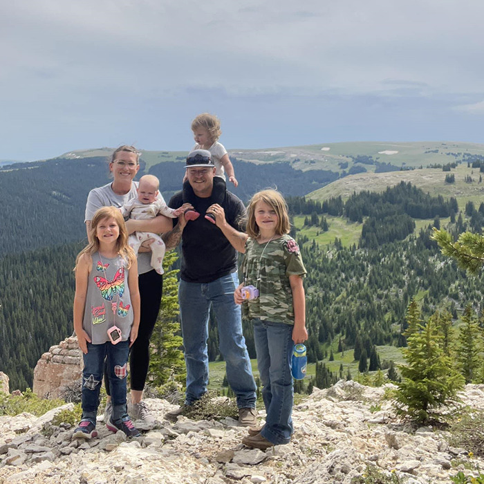 Family poses outdoors on a mountain with trees in the background; father stands with four daughters and mother.