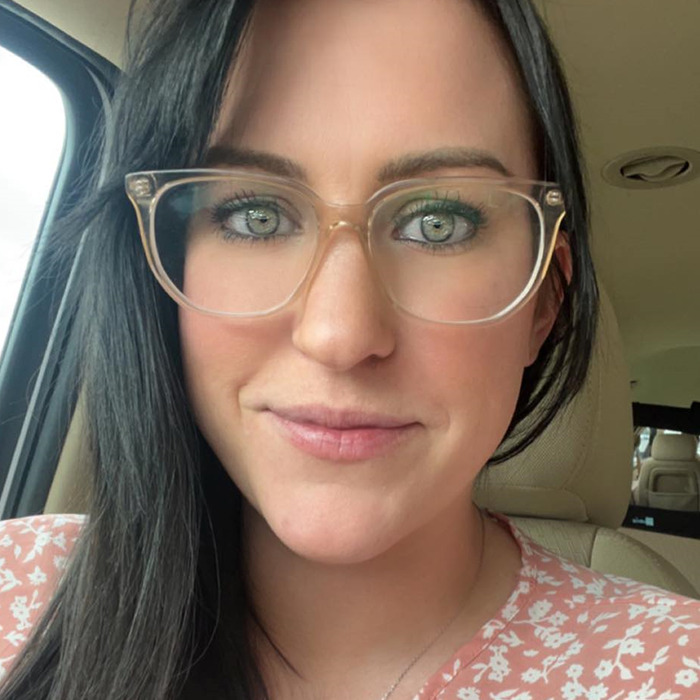 A woman with dark hair and glasses sits in a car, wearing a pink floral top.