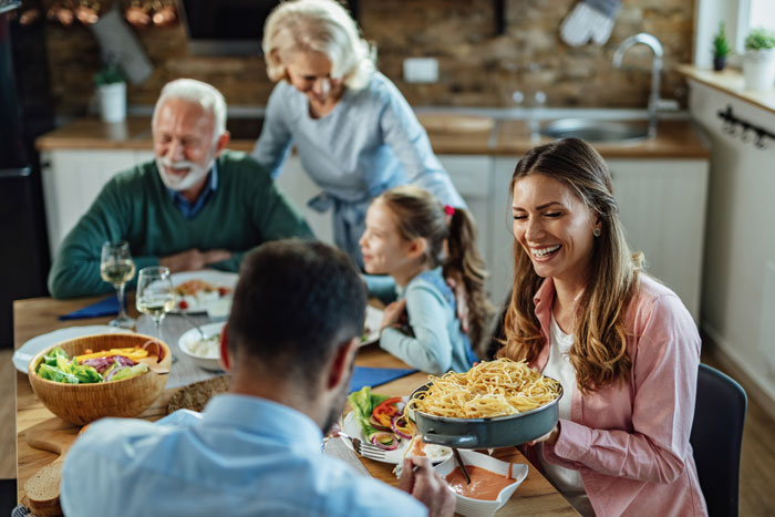 Happy family dinner, woman serving pasta, reflecting dynamic of mother-in-law being asked to leave for bringing uninvited guest.