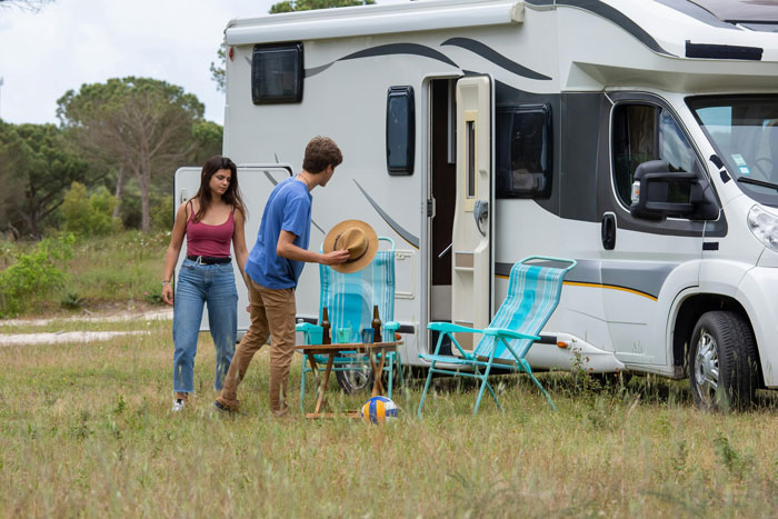 A couple stands by an RV; the man adjusts a hat on a camping chair while the woman observes, amidst a grassy field.