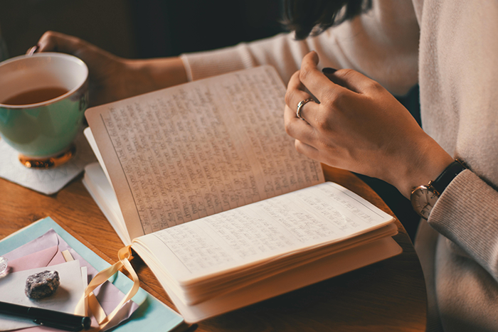 Hands holding an open journal with handwritten notes, along with a cup of tea nearby. Hands holding an open journal with handwritten notes, along with a cup of tea nearby.