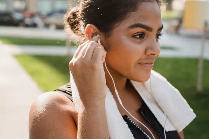 Woman at gym putting on headphones, towel around neck, preparing for workout in sunny outdoor setting.