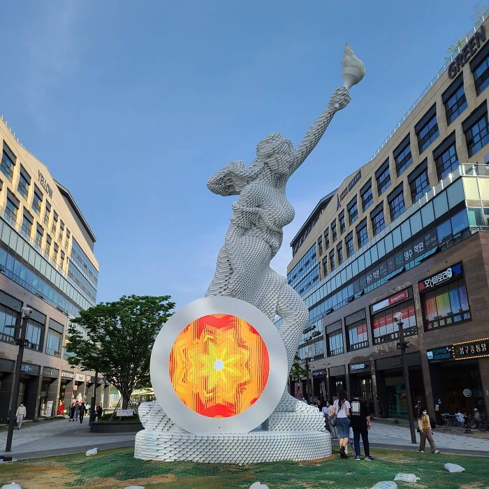 Metal pipe sculpture in an urban setting, featuring a figure with an illuminated shield and torch.