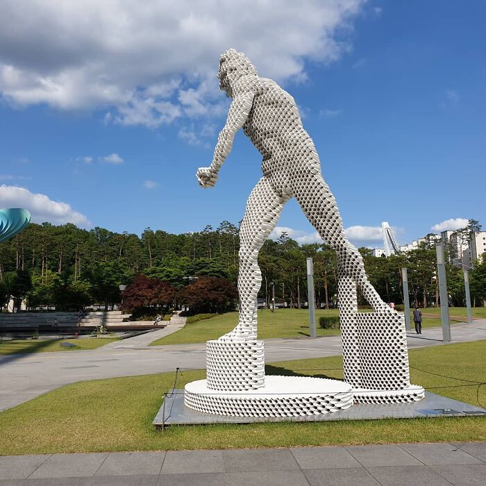 Metal pipe sculpture of a human figure by Yi Chul Hee in a park setting under a clear blue sky.