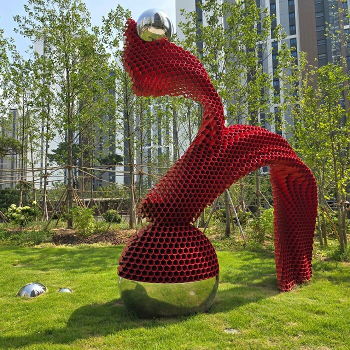 Metal pipe sculpture in a park, featuring a red abstract design with a shiny silver sphere, surrounded by trees.