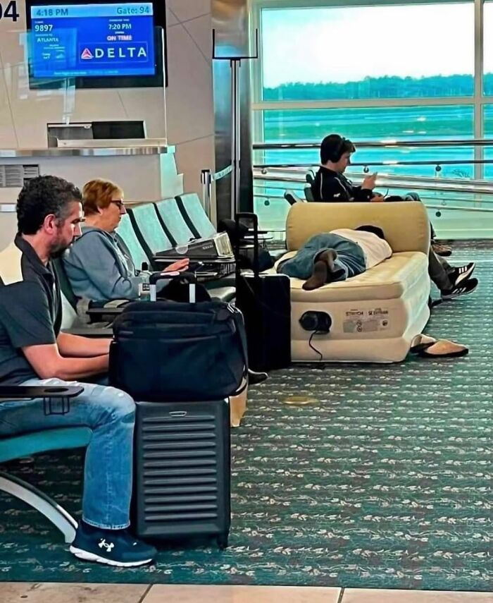 Personas esperando en el aeropuerto, una de ellas acostada en un colchón inflable, creando una escena divertida y confusa.