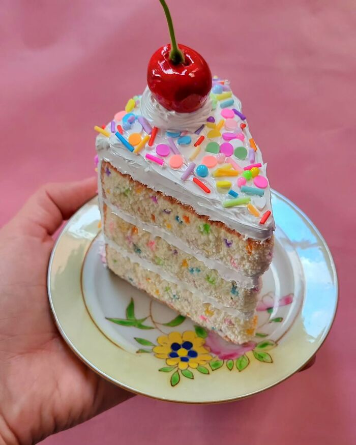 Hyper-realistic embroidered dessert resembling a slice of cake with a cherry on top, held on a decorative plate.