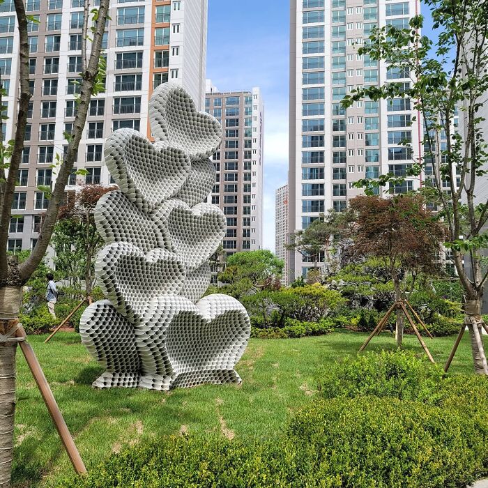 Metal pipe sculpture of stacked heart shapes by Yi Chul Hee displayed in a modern urban park setting.