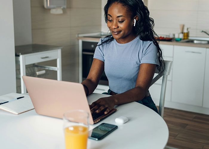 Young woman using laptop and wireless earbuds at kitchen table, demonstrating must-have life skills for internet users.