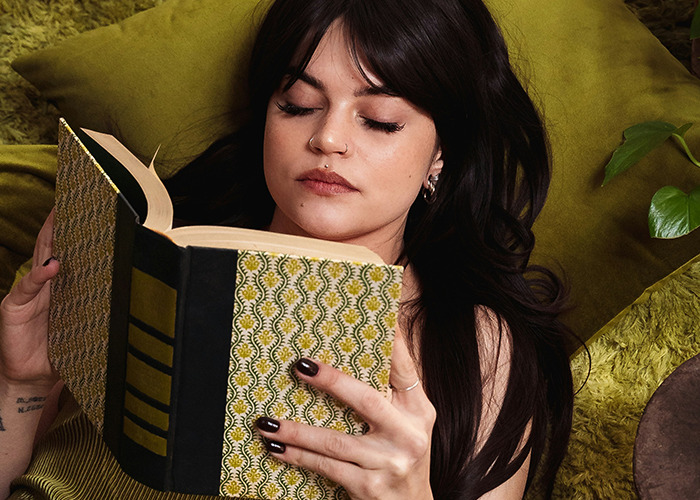 Young woman lying on green sofa, reading book about must-have life skills that nobody taught internet users.