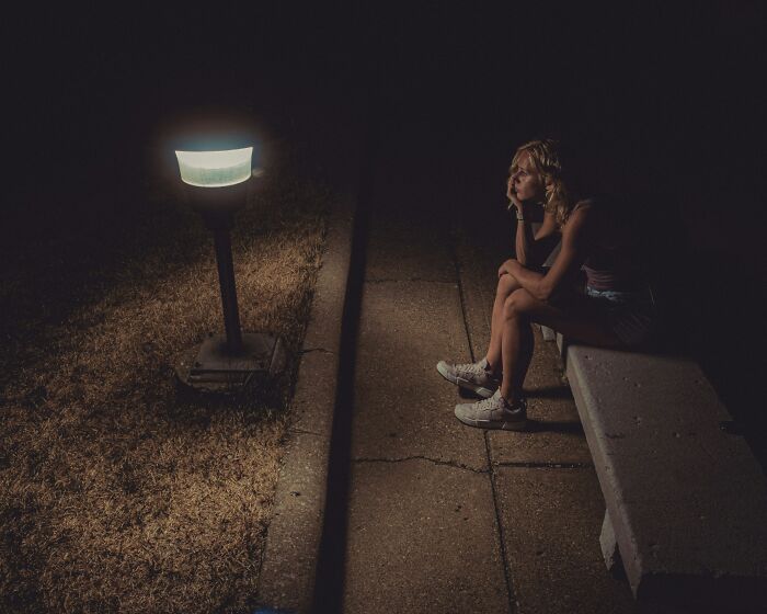 Person sitting alone on a bench at night, illuminated by a streetlamp, representing underrecognized traumatizing experiences.
