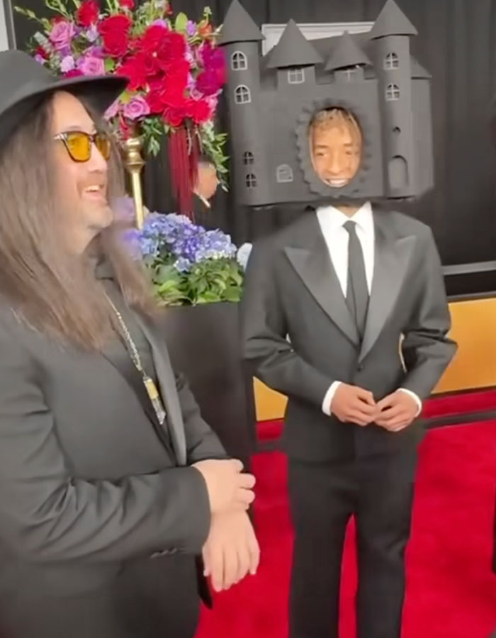 Two people in unique outfits on the red carpet, including a headpiece resembling a castle, creating a fashion statement. Two people in unique outfits on the red carpet, including a headpiece resembling a castle, creating a fashion statement.