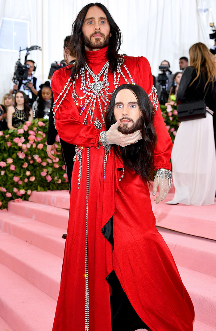 Celebrity in a red gown holding a replica of their own head at a high-profile event; an unforgettable and bold fashion statement.