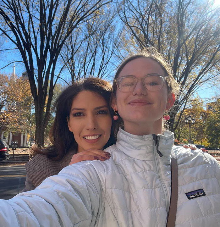 Two women smiling outdoors, one wearing glasses and a white jacket.