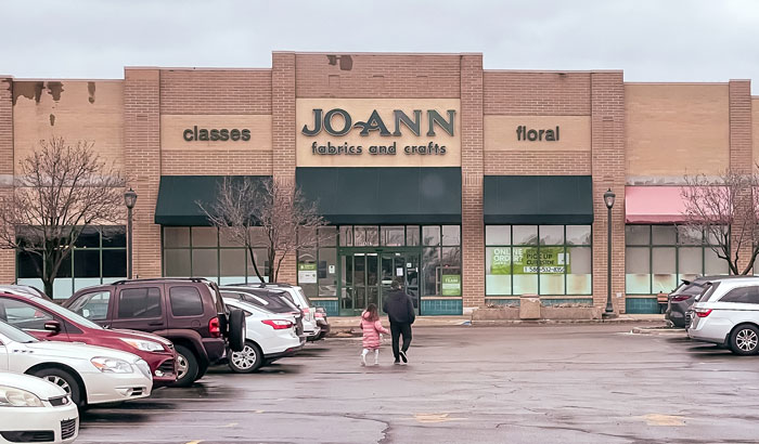 Joann Fabric and Craft store exterior, with people walking, showing brick facade and parking area.