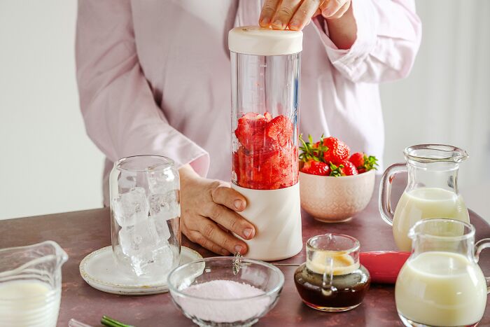 Person using a multipurpose blender with strawberries, surrounded by milk, coffee, and sugar on a kitchen counter.