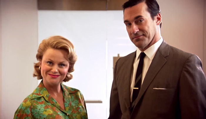 Jon Hamm and Amy Poehler in 1960s attire standing in an office setting.