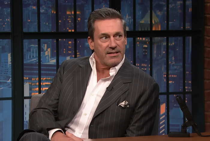 Jon Hamm in a pinstripe suit on a talk show set, addressing joke involving Amy Poehler.