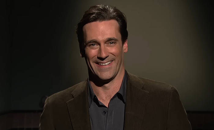 Jon Hamm smiling in a dimly lit setting, wearing a brown jacket.