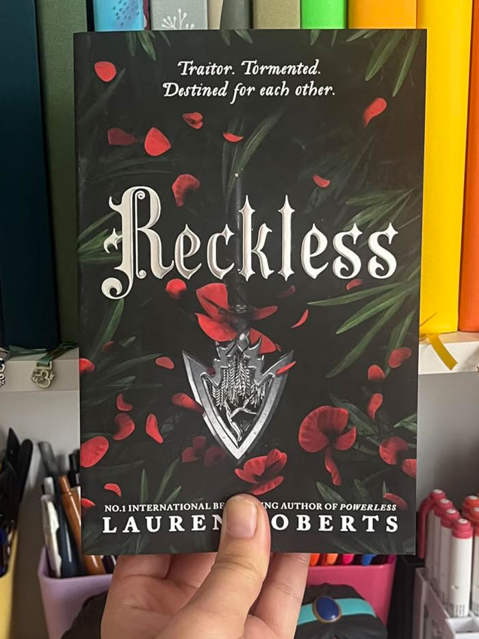 A person holding "Reckless" book with red petals on the cover, standing out against a colorful background.