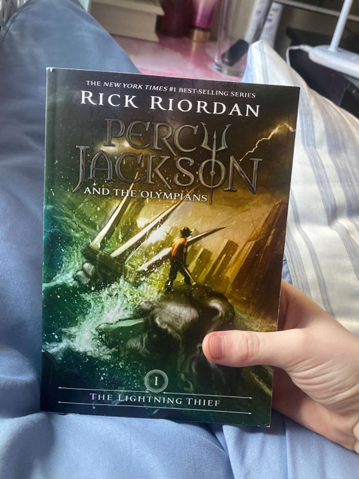 A hand holding the book "Percy Jackson and the Olympians" on a bed, showcasing one of the recommended book quotes.
