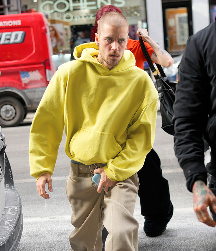 A person in a yellow hoodie and beige pants walks on a city street, holding a phone.