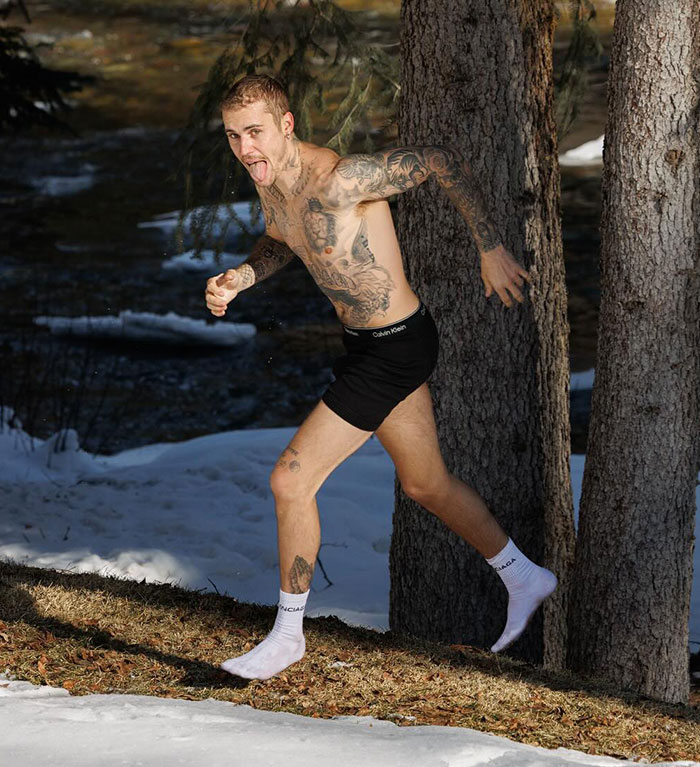 Man running in snow, shirtless and in black shorts, with tattoos visible, near a tree.