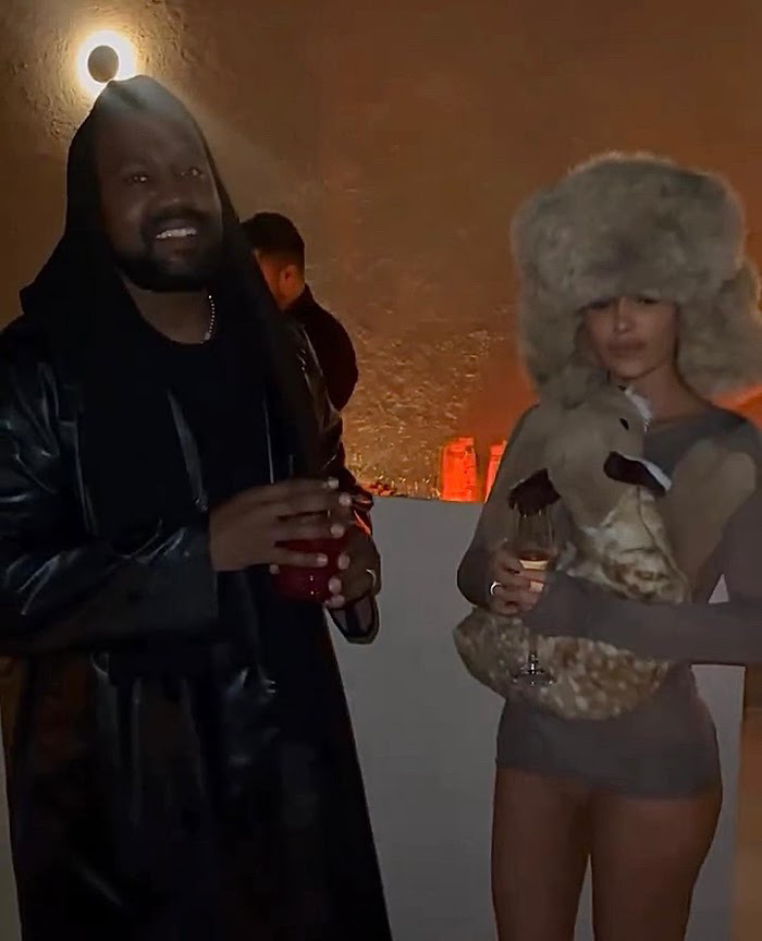 Bianca Censori and Kanye West at a dimly lit event, holding drinks and dressed stylishly. Bianca Censori and Kanye West at a dimly lit event, holding drinks and dressed stylishly.