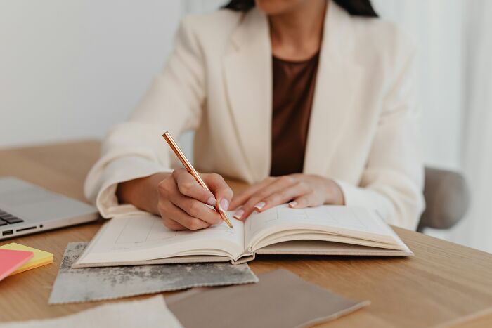 Woman in a white blazer writing in a notebook at a desk, capturing thoughts discreetly.