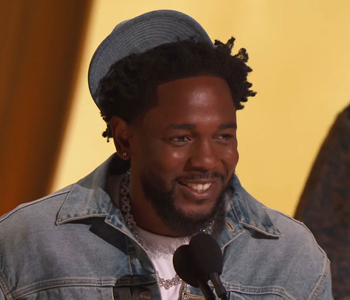 A man in a denim jacket and hat speaks at the 2025 Grammy Awards, smiling during a noteworthy moment.