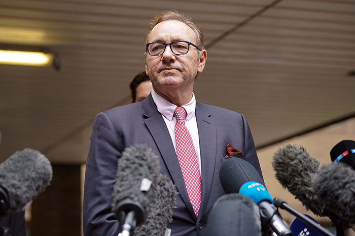 Kevin Spacey in a suit speaking to press, surrounded by microphones, related to Kevin Spacey claims.