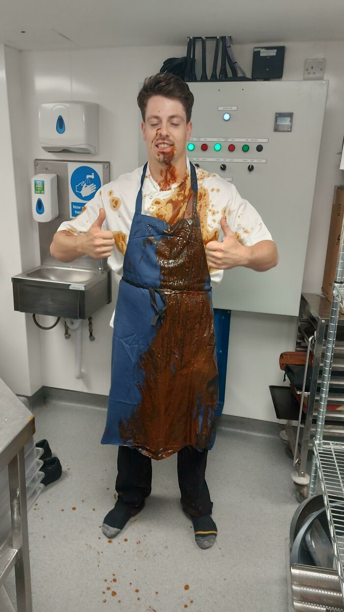 Worker in the food service industry with sauce all over their apron, giving thumbs up, in a kitchen setting.