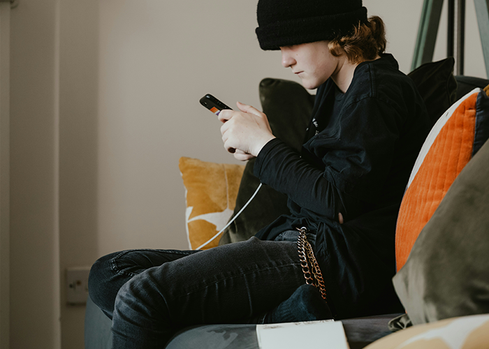 Teen in dark clothing sitting on a couch, focused on a smartphone, with pillows in the background.