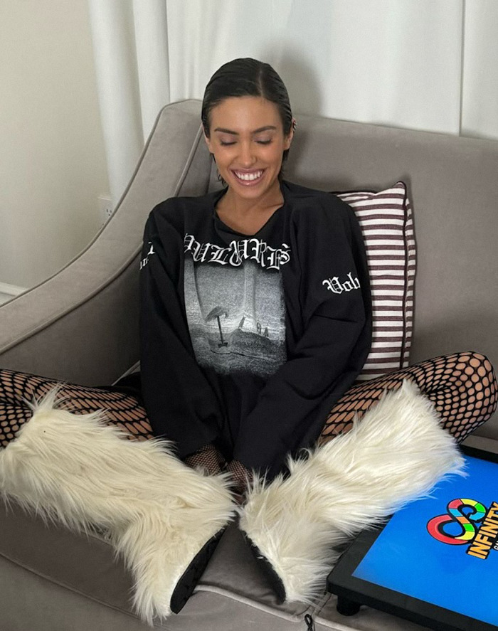 Bianca Censori in a black graphic T-shirt and furry boots sits cross-legged on a sofa, smiling. Bianca Censori in a black graphic T-shirt and furry boots sits cross-legged on a sofa, smiling.