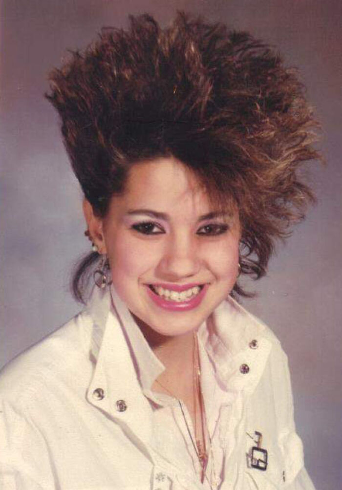 Woman with 1980s high hair, wearing a white jacket and earrings, smiling at the camera.