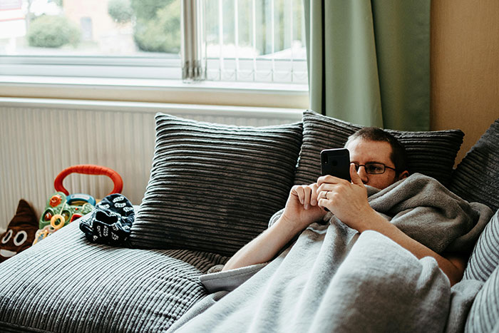 Man neglecting chores, lying on couch engrossed in phone, surrounded by unkempt living room with toys and pillows.