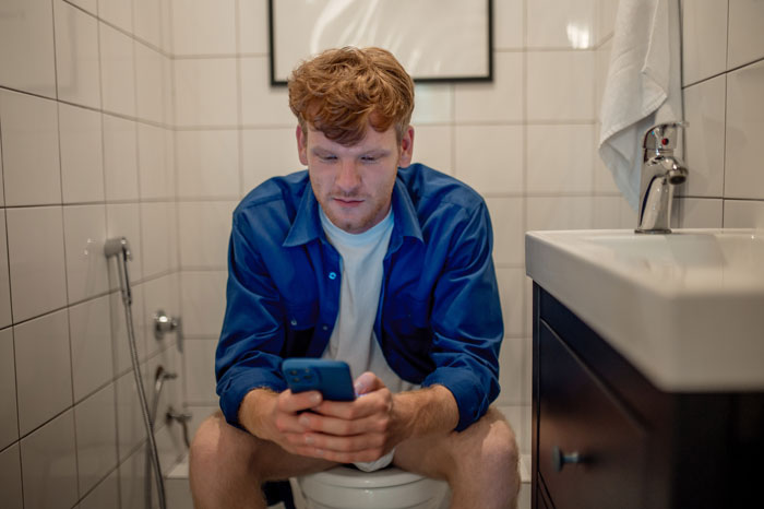Man in blue shirt using phone on toilet in a tiled bathroom, appearing focused.