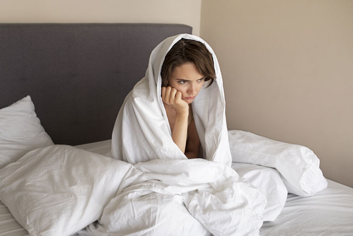 Woman wrapped in a duvet sitting on a bed, looking worried, reflecting a panic scenario in a hotel room.
