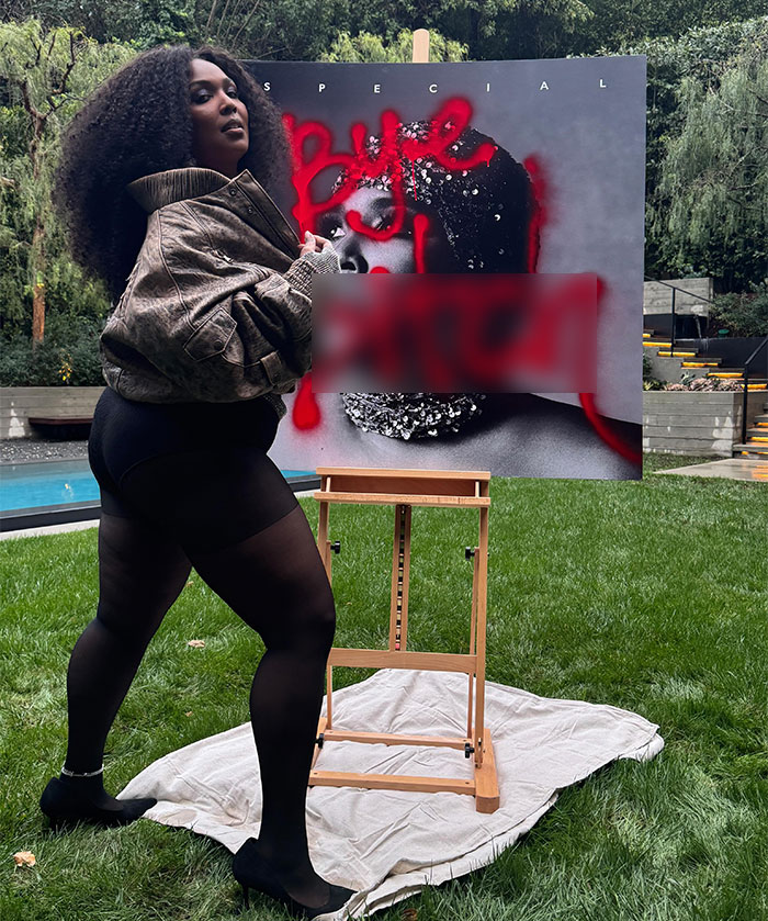 Person posing outdoors by a painted canvas, wearing a jacket and black shorts, showcasing a vibrant artistic expression.