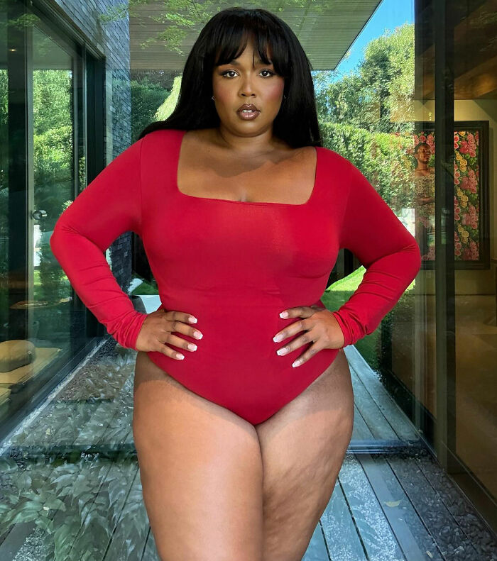 Person in a red bodysuit posing confidently indoors, hands on hips, with a glass window and greenery in the background.