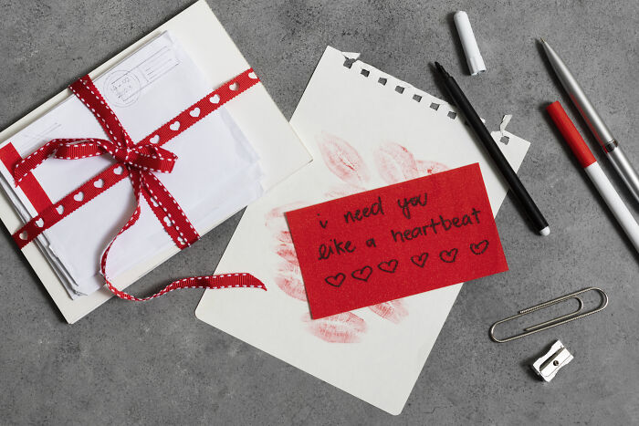 Valentine's gift idea with a red card, envelope tied with ribbon, pens, and a note saying "I need you like a heartbeat."