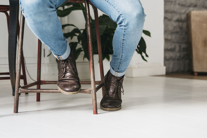 Casual footwear and jeans of everyday people sitting on stools, focus on leather boots, against a modern interior backdrop.