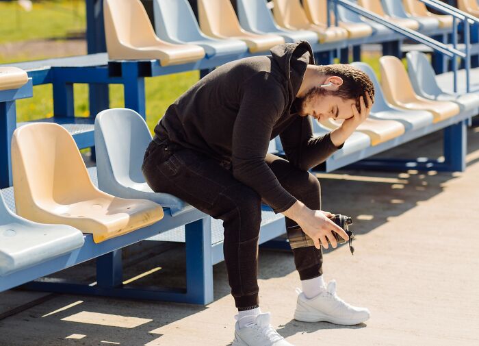 A man sitting in a stadium, looking downcast, clutching a water bottle, reflecting on life's harsh realities.