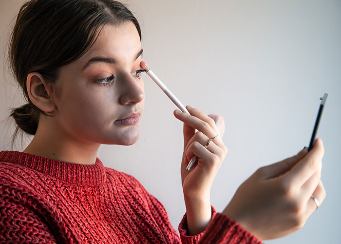 Woman applying makeup, holding a mirror, reflecting on coworker's audacity in a red sweater. Woman applying makeup, holding a mirror, reflecting on coworker's audacity in a red sweater.