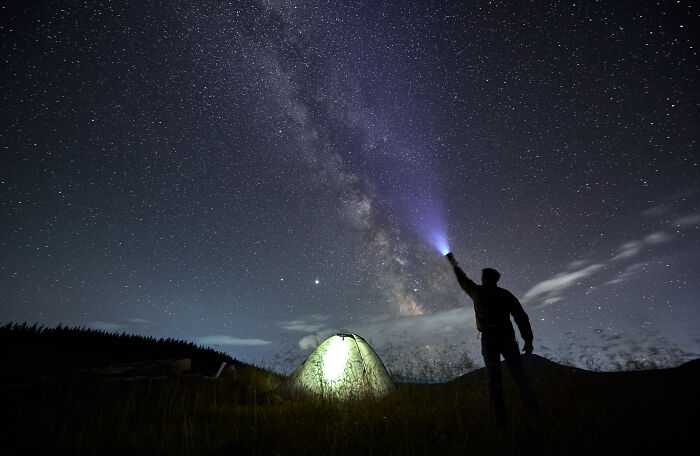 Person under starry sky with tent, shining flashlight at Milky Way—gift ideas for someone who has everything.