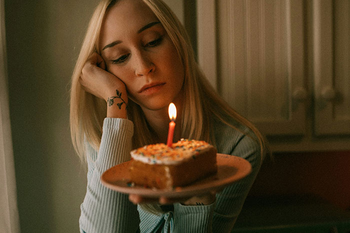 Woman holding a birthday cake with a candle, looking thoughtful in a dimly lit kitchen.