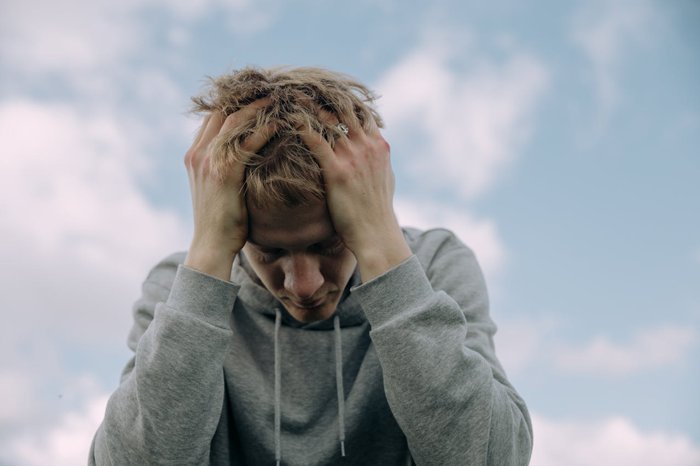 Man distressed, head in hands, wearing gray hoodie, with blue sky background, relating to relationship taking an odd turn.