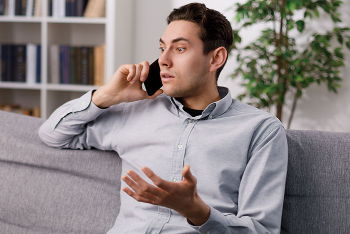 Man on phone discussing wedding and fiancée issues, sitting on a grey couch with bookshelf and plant in the background. Man on phone discussing wedding and fiancée issues, sitting on a grey couch with bookshelf and plant in the background.