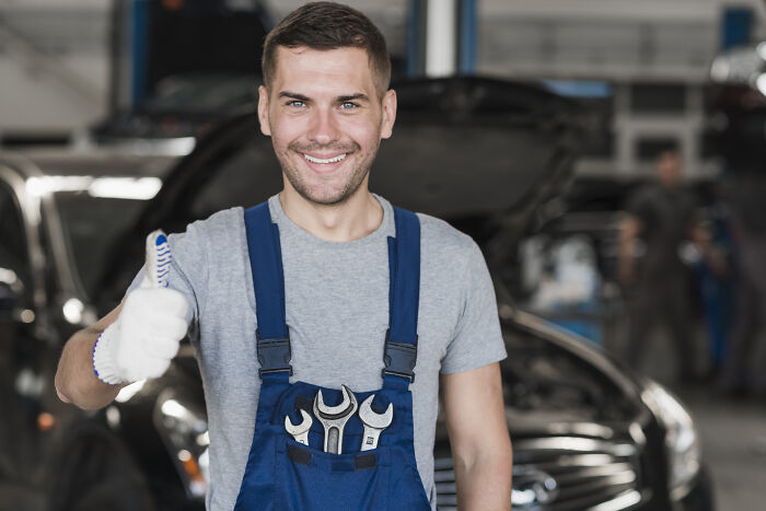 Mechanic in overalls smiling with thumbs up, warning about tire scam in an auto repair shop.