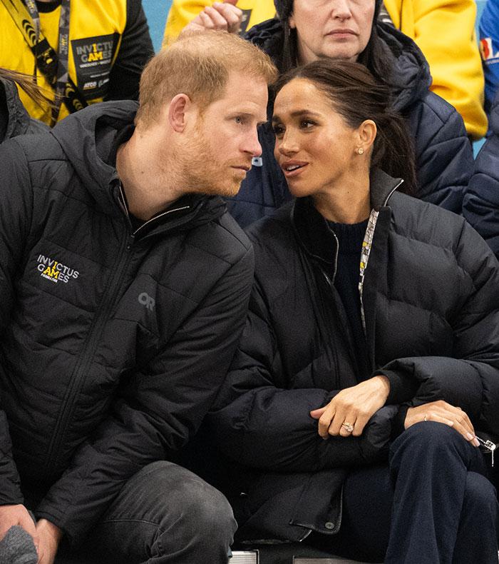 A couple seated, wearing black jackets at an event, engaging in a conversation. Hollywood is abuzz over Meghan Markle.